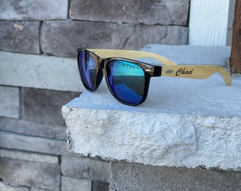 May include: Black sunglasses with blue mirrored lenses and a wooden frame. The frame is engraved with the name "Chad".