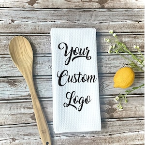 May include: White kitchen towel with a black script font that says "Your Custom Logo" on a wood background with a wooden spoon and a lemon.