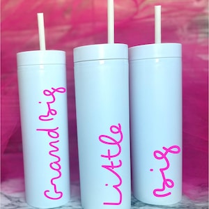 Puede incluir: Tres vasos cilíndricos blancos con tapas y pajitas blancas. Cada vaso presenta texto en cursiva de color rosa intenso. El vaso de la izquierda dice Grand Big, el central dice Little y el de la derecha dice Big en orientación vertical.