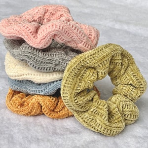 May include: A stack of five colorful scrunchies made of soft, textured fabric. The scrunchies are in shades of pink, gray, white, blue, and yellow.
