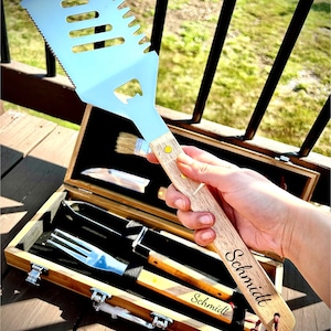 May include: A stainless steel barbecue spatula with a wooden handle, part of a grilling set. The handle is engraved with the name "Schmidt". The set includes a fork, knife, and brush, all stored in a wooden box.