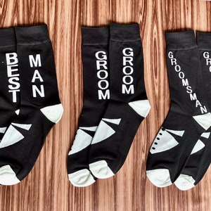 May include: Black socks with white text that reads "BEST MAN", "GROOM", and "GROOMSMAN". The socks have a white geometric design on the toe and heel. One pair of socks has a tuxedo graphic. Wedding party attire.