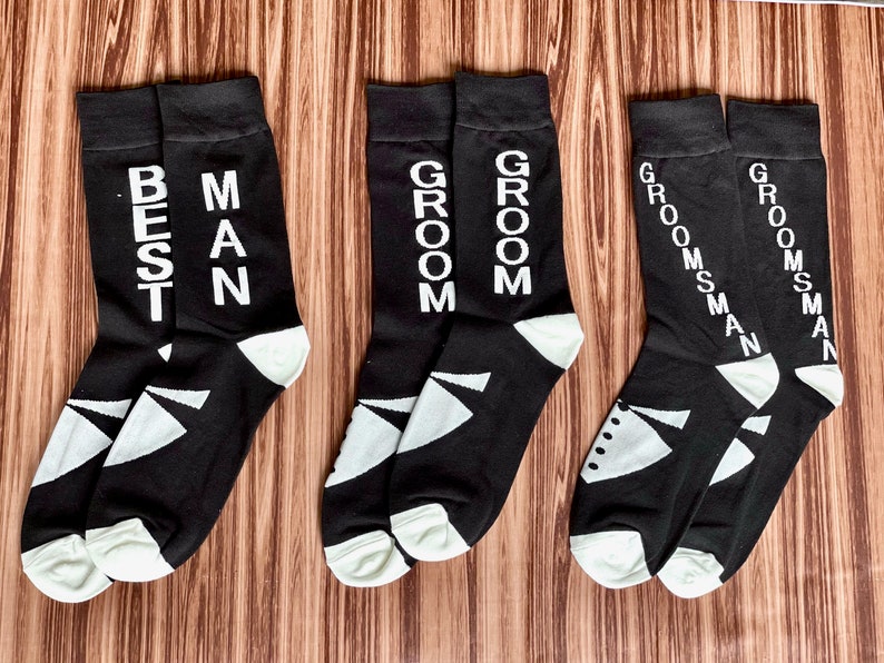 May include: Black socks with white text that reads "BEST MAN", "GROOM", and "GROOMSMAN". The socks have a white geometric design on the toe and heel. One pair of socks has a tuxedo graphic. Wedding party attire.