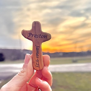 May include: A wooden cross with the name "Preston" engraved on it, along with the text "James 9:23". The cross is held up against a sunset backdrop, showcasing its natural wood grain and unique shape.