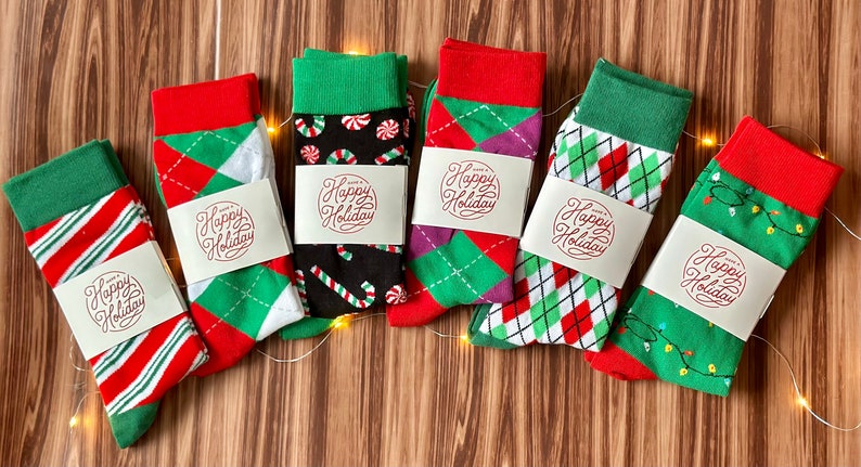 May include: Six pairs of colorful Christmas socks with green, red, and white patterns. Each sock has a white tag with the text "Happy Holiday" in red.