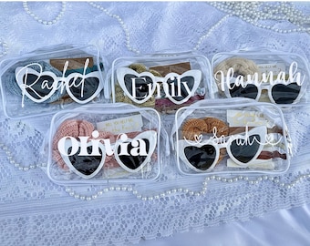 May include: Four clear plastic bags with zipper closures, each containing a pair of heart-shaped sunglasses with a white name written on them. The names are: Rachel, Emily, Hannah, and Olivia. Each bag also contains other small items, such as hair ties and cards.