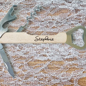 May include: A multi-tool with a light wood handle, featuring a corkscrew, bottle opener, and knife. The name "Stephanie" is written on the handle in black script. The tool is made of stainless steel and wood.