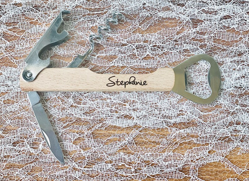 May include: A multi-tool with a light wood handle, featuring a corkscrew, bottle opener, and knife. The name "Stephanie" is written on the handle in black script. The tool is made of stainless steel and wood.