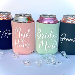 May include: Four beverage insulators with different colors and text. The insulators are navy blue, pink, light green, and black. The text on the insulators reads "Best Man", "Maid of Honor", "Bridesmaid", and "Groomsman".