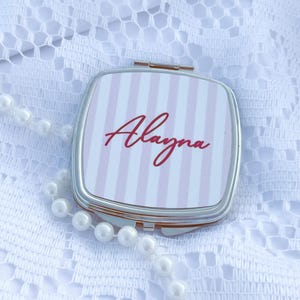 May include: A square silver compact mirror featuring a light pink and white vertical striped pattern on the lid. The name Alayna is printed in a dark red cursive script across the center of the striped design on the front of the mirror.
