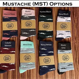 May include: A collection of eight pairs of socks with mustache patterns in various color combinations, including mauve/navy, blush/charcoal, hunter/black, and navy/blush. Each pair is wrapped with a brown paper band that reads "SUIT UP". The text "MUSTACHE (MST) OPTIONS" is at the top.