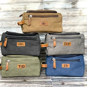 May include: Four canvas toiletry bags with leather accents and personalized initials. The bags are stacked on top of each other. The top bag is brown, the second bag is gray, the third bag is green, and the bottom bag is blue.