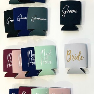 May include: Assortment of colorful can coolers with various wedding party titles printed in white and gold lettering. The titles include "Best Man", "Groom", "Groomsman", "Maid of Honor", "Bride", and "Bridesmaid".