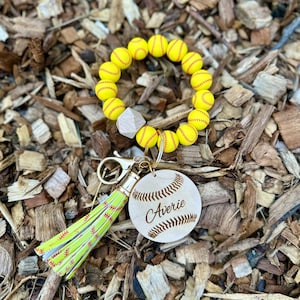 May include: A softball-themed keychain with a yellow beaded bracelet, a wooden charm engraved with "Averie", and a green and yellow tassel. The charm is shaped like a softball with stitched details.