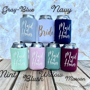 May include: Set of seven beverage insulators with different colors and text. The colors are gray-blue, navy, mint, blush, willow, and maroon. The text on the insulators reads "Maid of Honor" and "Bride".