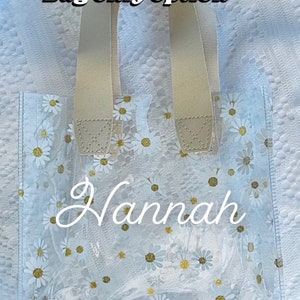 Może przedstawiać: Przezroczysta torba z tworzywa sztucznego z nadrukiem białych stokrotek i beżowymi uchwytami. Na torbie widnieje napis "Hannah".