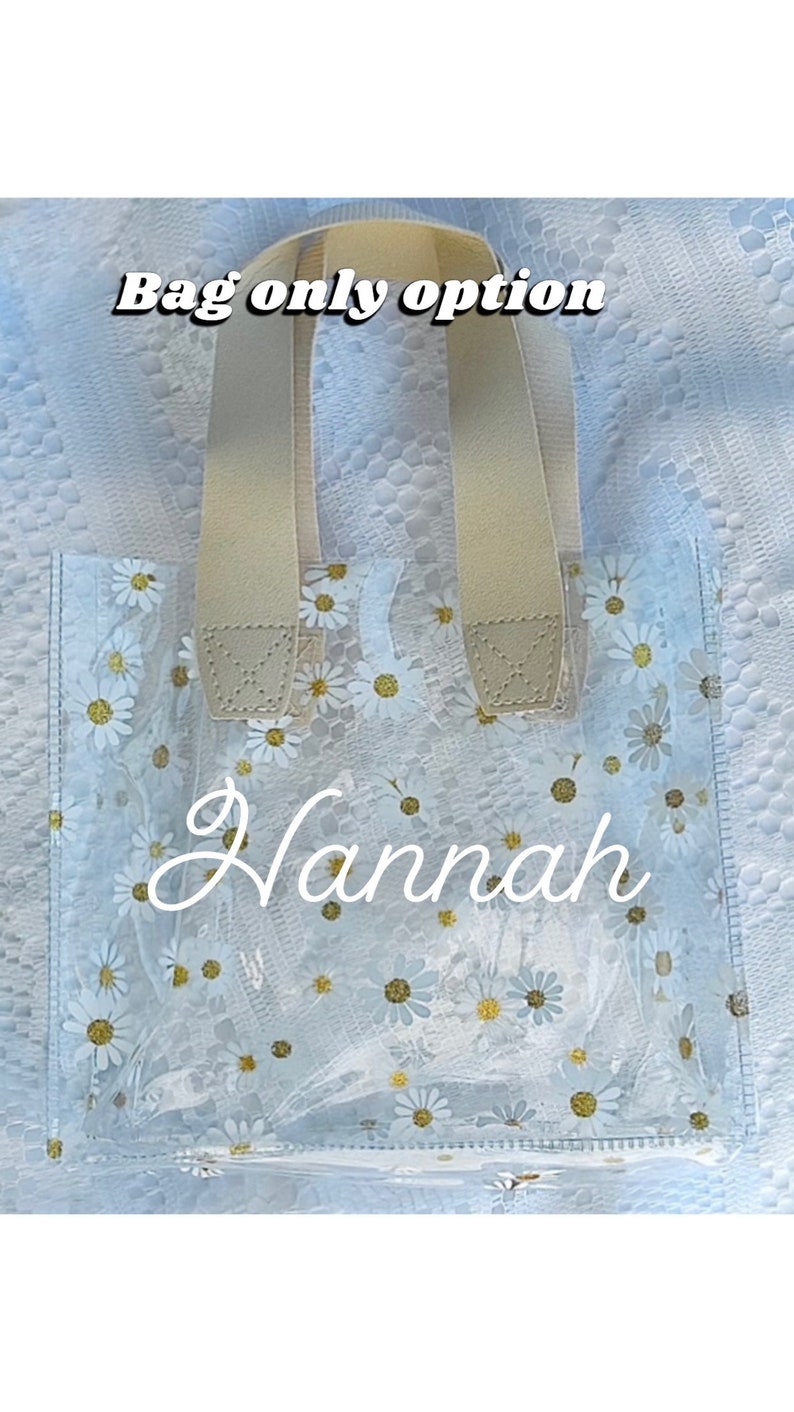 Może przedstawiać: Przezroczysta torba z tworzywa sztucznego z nadrukiem białych stokrotek i beżowymi uchwytami. Na torbie widnieje napis "Hannah".