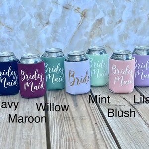 May include: Six different colored can coolers with the text "Brides Maid" or "Bride" printed in white. The colors are navy, maroon, green, white, pink, and lilac.