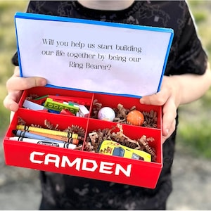 May include: A red box with the name "CAMDEN" on the front. The box is open and contains a variety of small toys, including crayons, a toy car, and a small ball. The box also has a note inside that says "Will you help us start building our life together by being our Ring Bearer?"