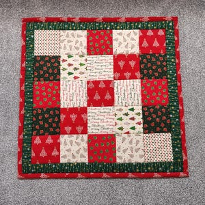 May include: A square Christmas placemat with a patchwork design. The placemat features red, green, and white squares, some with Christmas tree patterns and others with the text "Merry Christmas".