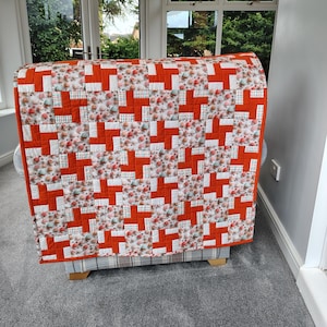 May include: A handmade quilt draped over a chair. The quilt features a repeating pattern of orange and white crosses on a floral background. The quilt's border is a solid orange color.
