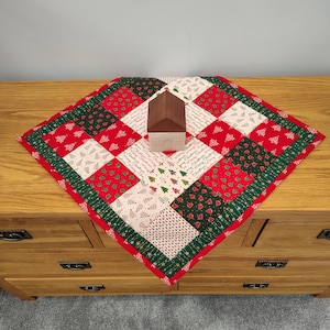 May include: A festive Christmas table runner with a patchwork design in red, green, and white. The runner features Christmas tree and holiday message patterns. A small wooden house sits on top of the runner, adding a decorative touch.