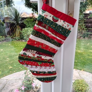 May include: A festive Christmas stocking, suspended from a door handle. The stocking is crafted from various fabric strips in red, green, and white. It showcases Christmas motifs such as stars, baubles, and snowmen. The top section is red, with the words "Merry Christmas".