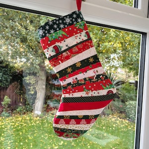 May include: A festive Christmas stocking hanging from a window. The stocking features a patchwork design with red, white, and green fabric strips adorned with Christmas motifs. The background shows a garden with green grass and trees.
