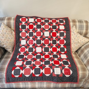 May include: A handmade quilt featuring a geometric design of red, white, and black squares. The quilt is placed on a plaid sofa. The border is black with a red dotted trim. A decorative item for the home.