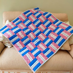 May include: A pink and blue patchwork quilt with a woven pattern. The quilt is made of fabric with a small white and blue floral print.