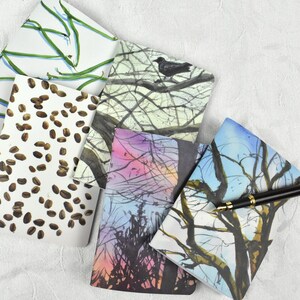 May include: Five small notebooks with different watercolor designs. The notebooks have covers with images of coffee beans, tree branches, and a bird on a branch. A black pen with gold accents is lying on top of the notebooks.