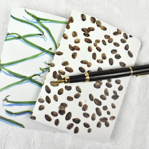 May include: Two small notebooks with different designs. One notebook has a white background with green lines and the other has a white background with brown coffee beans. A black and gold fountain pen is lying on top of the notebooks.