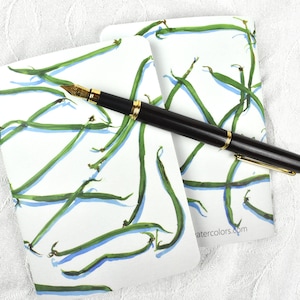May include: Two white notecards with a watercolor illustration of green beans, with a black and gold fountain pen resting on top. The notecards have a rounded top and are on a white textured surface.