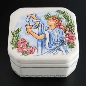 Könnte beinhalten: Eine weiße Keramikdose mit floralem Design. Der Deckel zeigt eine Frau, die Wasser aus einem Krug in eine Tasse gießt. Die Frau trägt ein blaues Kleid und einen Kranz aus Blumen auf dem Kopf. Die Dose ist mit grünen Blättern und rosa Blumen verziert.