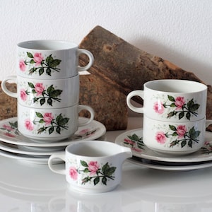 Könnte beinhalten: Ein Set aus vier weißen Teetassen mit rosafarbenen Rosenblütenmustern, die übereinander gestapelt sind. Die Teetassen stehen auf einem weißen Unterteller mit passendem Blumenmuster. Im Vordergrund befindet sich ein kleiner weißer Milchkännchen mit passendem Blumenmuster.