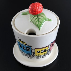 May include: White ceramic sugar bowl with a lid featuring a red strawberry and green leaf handle. The bowl has a colorful train design around the base and sits on a matching saucer. A charming kitchenware item.