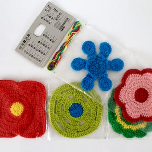 Op de afbeelding: Een verzameling kleurrijke gehaakte bloemvormen in een transparante plastic verpakking. De bloemen zijn rood, roze, blauw en groen met gele en blauwe centra. Een klein pakket met instructies en kleurrijk garen is ook inbegrepen.