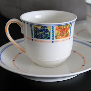 Può includere: Un set da tè bianco con tazza e piattino con un motivo floreale in tonalità di blu, giallo e arancio. La tazza ha una forma arrotondata e un manico. Il piattino ha un bordo ondulato.