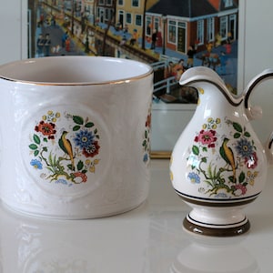 Könnte beinhalten: Ein weißer Keramik-Pflanzer und Krug mit floralem und Vogelmotiv. Der Pflanzer hat einen goldenen Rand und der Krug einen braunen Rand.