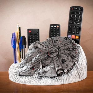 Puede incluir: Un organizador de escritorio gris y texturizado con forma de la nave espacial Millennium Falcon. La estructura cuenta con múltiples ranuras que sostienen varios bolígrafos, lápices y tres controles remotos negros de diferentes tamaños. La base tiene un aspecto rocoso.