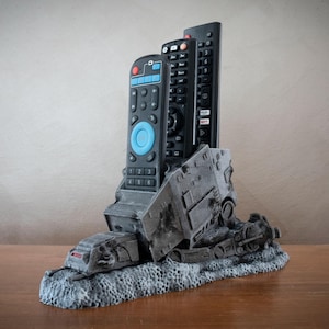 May include: A grey, 3D printed remote control holder shaped like the Death Star from Star Wars. The holder has three slots for remotes and is sitting on a wooden surface.