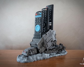 Può includere: Un porta telecomando grigio stampato in 3D a forma di Morte Nera di Star Wars. Il porta telecomando ha tre slot per i telecomandi ed è posizionato su una superficie di legno.