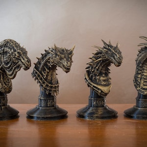 May include: Four detailed dragon head busts, each with a different design and color scheme. The busts are mounted on black bases with gold accents. The bases are labeled with the names "Thyr", "Caga", "Mick", and "Syrax".