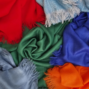 Pode incluir: Uma coleção de cachecóis retangulares macios em cores sólidas vibrantes, incluindo vermelho, verde floresta, azul royal, laranja brilhante, cinza claro e um azul claro texturizado, todos com franjas delicadas nas extremidades curtas do tecido.