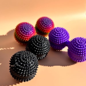 May include: A selection of spiky massage balls in different colours. The balls are black, purple, and a mix of red, purple, and green. They are arranged on a peach-coloured surface.