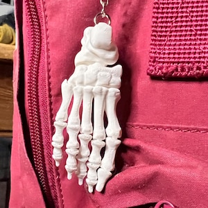 Puede incluir: Un llavero de mano esquelética blanca cuelga de una cadena plateada. El llavero es una réplica detallada de una mano humana, con huesos individuales visibles. El fondo es una tela rosa-roja, con una cremallera y una correa.