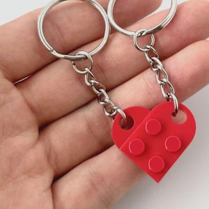May include: A pair of keychains featuring a red heart design made of interlocking blocks. Each keychain has a silver chain and a metal ring. The heart is divided into two halves, each with three circular studs.