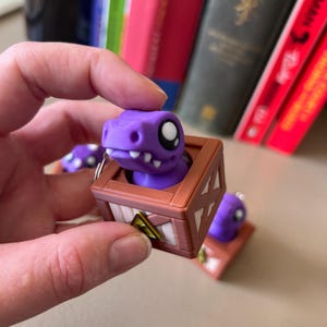 Könnte beinhalten: Eine lila Dinosaurierfigur in einer braunen Holzkiste, mit einem weißen Auge und einem gelb-schwarzen Warnschild. Die Figur wird von einer Hand gehalten, mit anderen Figuren und Büchern im Hintergrund.