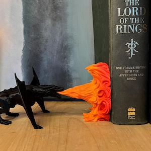 May include: A black dragon-shaped bookend with orange flames, positioned next to a stack of books, including "The Lord of the Rings." The dragon appears to be breathing fire, adding a fantasy element to the bookshelf.