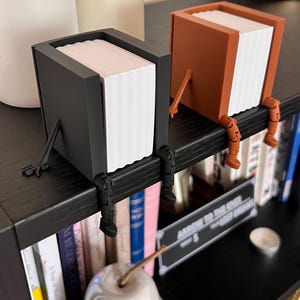 Puede incluir: Dos originales sujetalibros, uno negro y otro marrón, cada uno con forma de caja con una pila de papel blanco en su interior. Tienen piernas y pies naranjas, como si estuvieran sentados en una estantería negra. Un objeto de cerámica con forma de pera en primer plano.